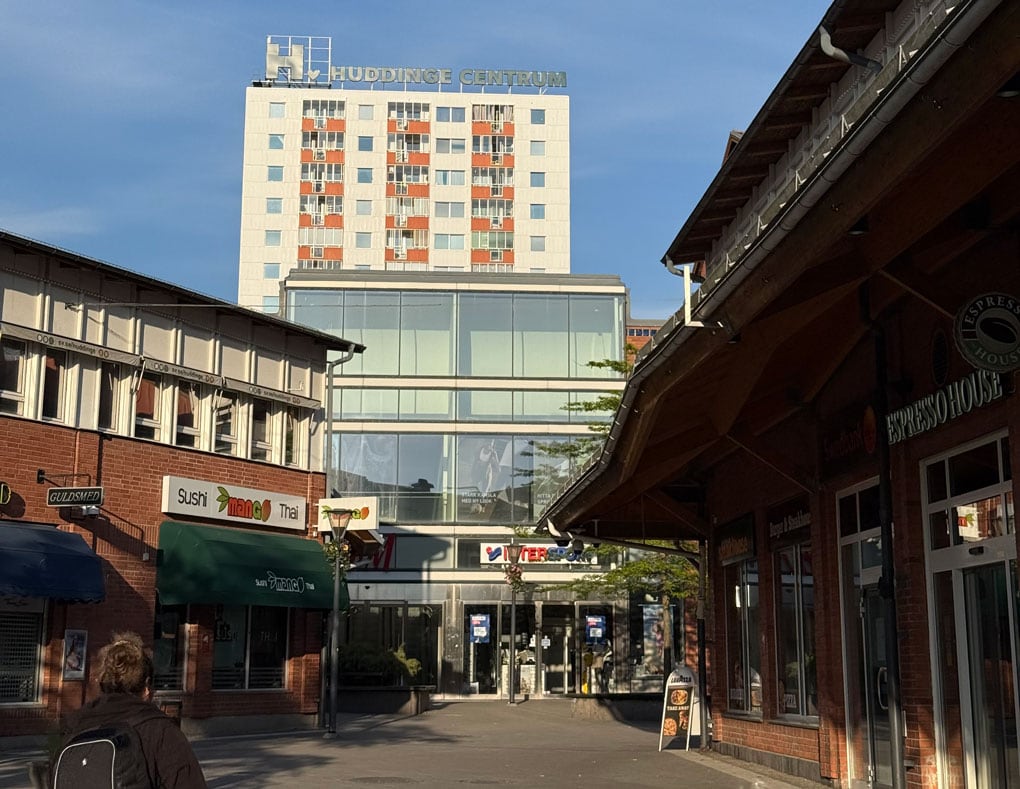 Huddinge centrum med butiker och restauranger längs gågatan och höghuset med skylten Huddinge Centrum i bakgrunden.