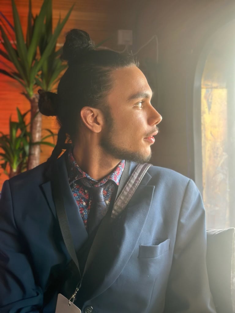 Adam Hassad i blå kostym och mönstrad skjorta sitter inomhus och blickar ut genom ett fönster, med varmt solljus över ansiktet.