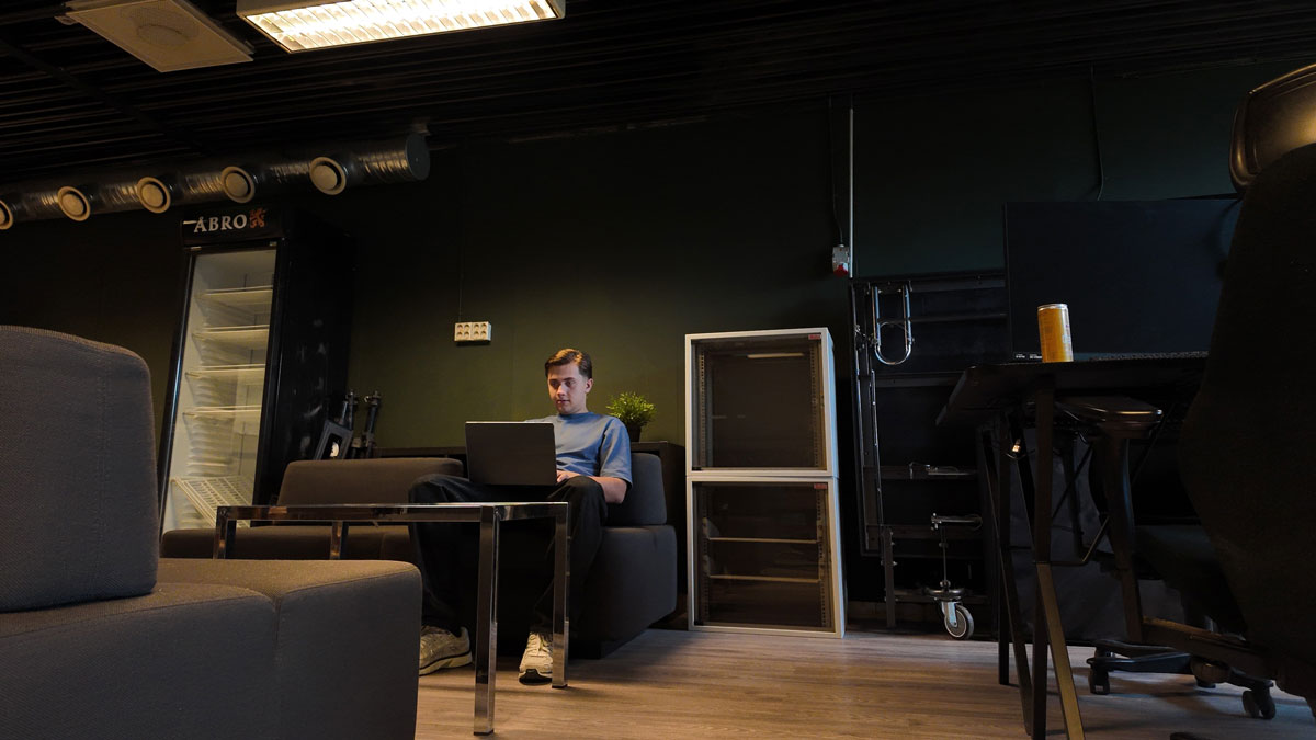 En person sitter i en soffa och arbetar vid en bärbar dator i en mörk lokal med sittgrupper och bord.