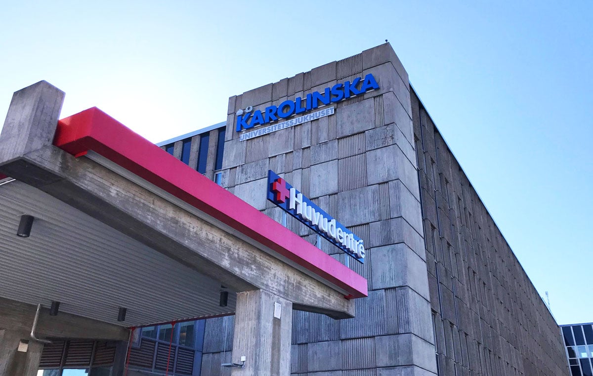 Karolinska Universitetssjukhusets grå betongbyggnad i Huddinge, med huvudentréns röda tak i förgrunden och blå sjukhusskylt på fasaden.