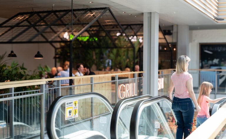 Interiör i Skogås centrum med rulltrappor, restaurang och besökare som rör sig genom köpcentrumet.