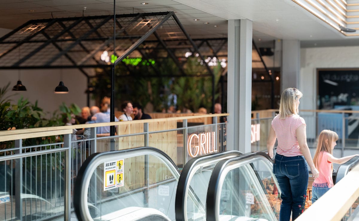 Interiör i Skogås centrum med rulltrappor, restaurang och besökare som rör sig genom köpcentrumet.