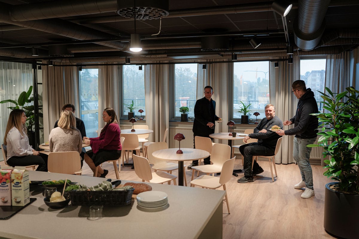 Personer samtalar och äter tillsammans vid små bord i en ljus coworkingmiljö med stora fönster och gröna växter.